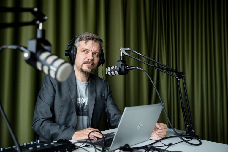 Iltalehden julkaisupäällikkö Mikko Räsänen istuu paksun verhon edessä podcast-studiossa kannettavan tietokoneen ja mikrofonien ääressä.