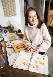 Muhkeaan neuleeseen puheutunut Riikka Haahti istuu työpöydän ääressä. Hän sormeilee muistikirjaa, johon on sivelty maalisävyjä ja kirjattu muistiinpanoja. Edessään hänellä on pieni, keskeneräinen ikonimaalaus. Aiheena on madonna ja lapsi.