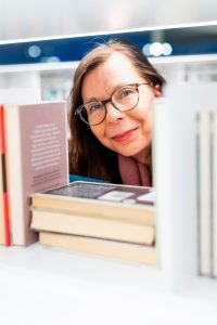 Tummahiuksinen ja silmälasipäinen Maarit Piippo kurkistaa hymyillen kuvaajaa hyllytettyjen kirjojen lomasta.