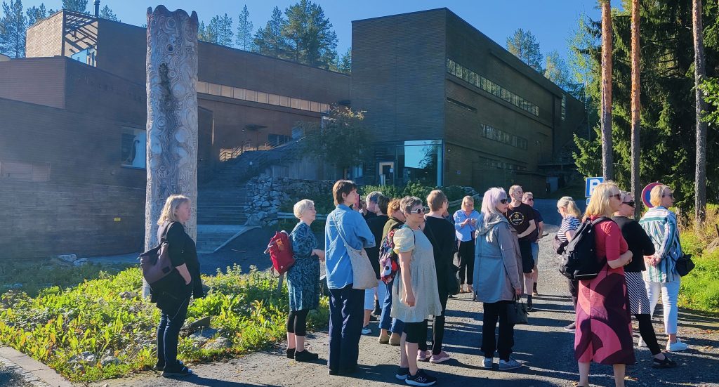 Kansantaiteenkeskus levittäytyy Kaustisella kallioiseen rinteeseen. yli 3600 neliötä päällystää puinen vaakalaudoitus ja rakennuksen keskeltä johtavat kiviset portaat Puhkion kallion huipulle asti. Keskuksen edessä ryhmämme levittäytyy kuulemaan rakennuksen historiasta ja tarkoituksista.