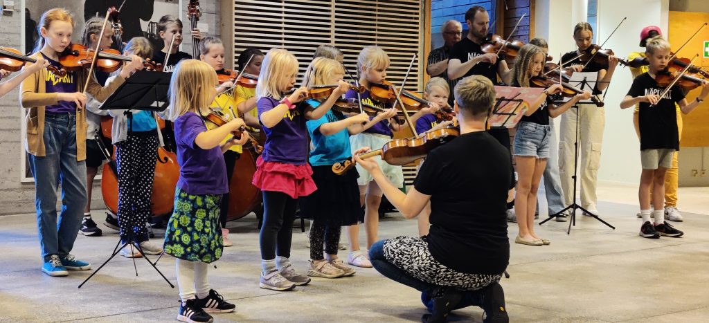 Pienen pienet viulistit etualalla saavat ohjausta omalla tasollaan. Ohjaaja vastapäätä koukistuu heidän eteensä näyttämään mallia. Kappaleita opetellaan alkuun ryhmässä. Takarivissä osataan jo lukea nuotteja, ja kappaleet sointuvat pian korvakuulolta. 