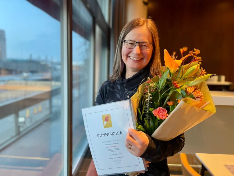 Hymyilevä Tarja Hirvasnoro poseeraa kameralle sylissään Salli-diplomi ja onnittelukukat hotelli Presidentin ikkunan ääreellä.