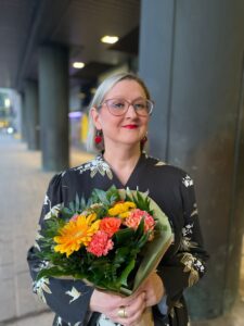 Heta Saxell poseeraa kameralle hotelli Presidentin paasien vierellä, kädessään onnittelukukat.