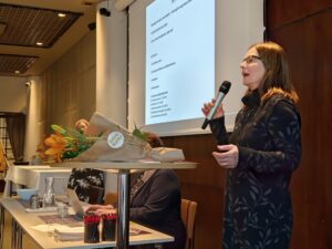 Tarja Hirvasnoro pitää puhetta Presidentin Sylvi-salissa, kukat nojaavat hänen edessään korkealla pöydällä.